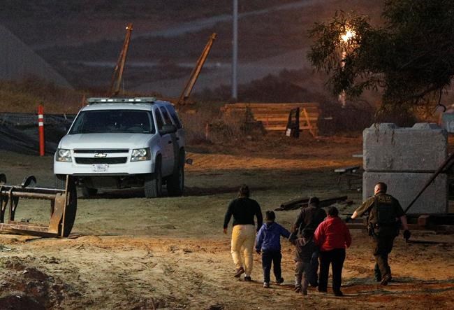 FILE - In this Dec. 3, 2018, file photo, migrants are escorted by a U.S. Border Patrol agent as they are detained after climbing over the border wall from Playas de Tijuana, Mexico, to San Ysidro, Calif. The Trump administration expects to launch a policy as early as Friday, Jan. 25, that forces people seeking asylum to wait in Mexico while their cases wind through U.S. courts, an official said, marking one of the most significant changes to the immigration system of Donald Trump's presidency. (AP Photo/Rebecca Blackwell, File)