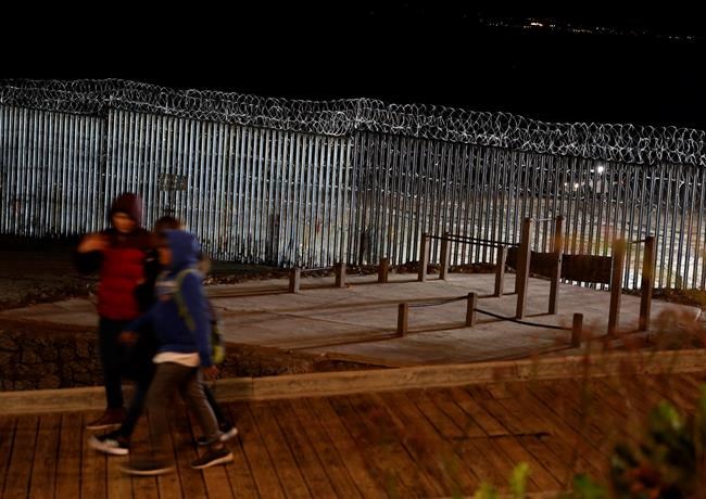 Floodlights from the U.S, illuminate the border wall, topped with razor wire, as a people pass Monday, Jan. 7, 2019, at the beach in Tijuana, Mexico. With no breakthrough in sight, President Donald Trump will argue his case to the nation Tuesday night that a