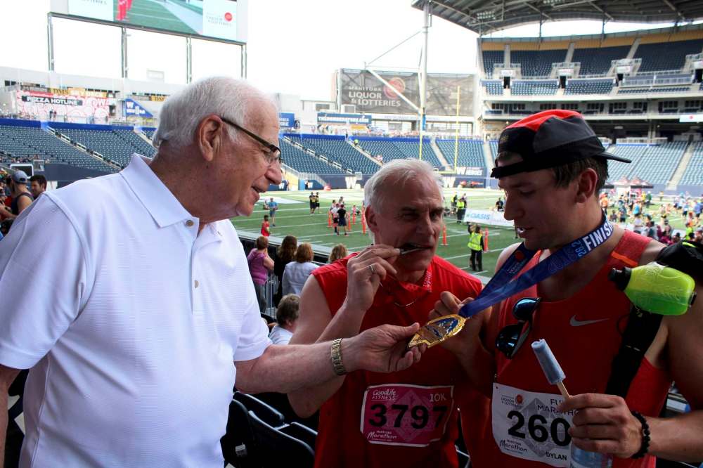 Arie Bronk, 81, touches his grandson Dylan Bronk's Manitoba Marathon medal Sunday as Bob Bronk, 58, bites his 10-km medal.