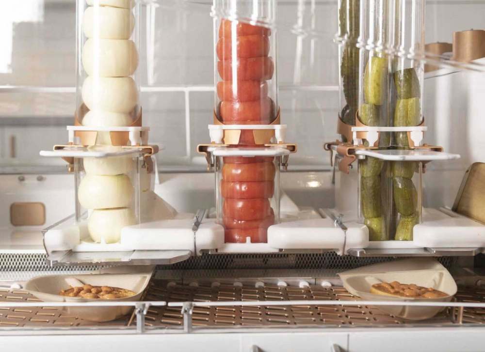 Burgers are topped with condiments while moving through the machine at the Creator storefront in San Francisco, California, U.S., on Monday, June 18, 2018. On June 27, the world's first robot-crafted burger will roll off a conveyor belt in San Francisco and into the hands of the public. The product, from Bay Area-based Creator, a culinary robotics company, is assembled and cooked in a machine that contains 20 computers, 350 sensors, and 50 actuator mechanisms. Photographer: Cayce Clifford/Bloomberg