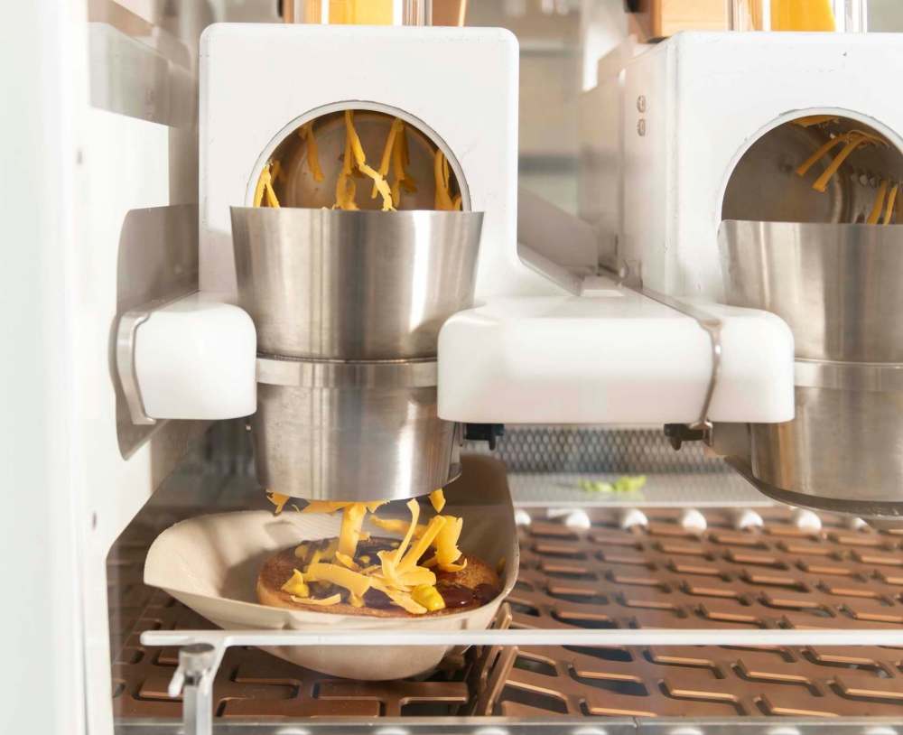 The machine shreds cheese onto a bun at the Creator storefront in San Francisco, California, U.S., on Monday, June 18, 2018. On June 27, the world’s first robot-crafted burger will roll off a conveyor belt in San Francisco and into the hands of the public. The product, from Bay Area-based Creator, a culinary robotics company, is assembled and cooked in a machine that contains 20 computers, 350 sensors, and 50 actuator mechanisms. Photographer: Cayce Clifford/Bloomberg