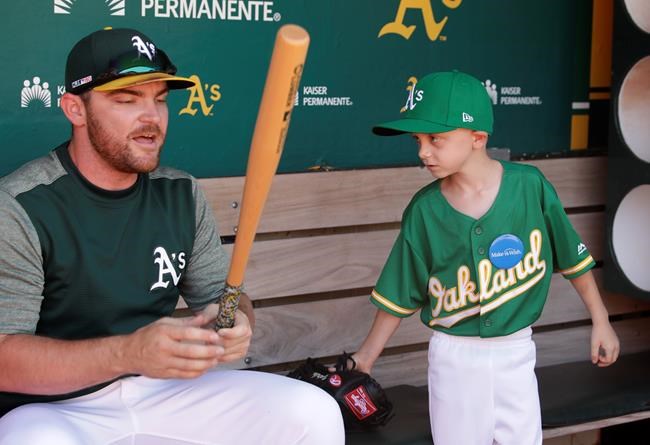 Eight-year-old August Wold, right, waits for Oakland Athletics' Liam Hendriks to finish taping his bat in the dugout prior to a baseball game against the Texas Rangers, Saturday, July 27, 2019, in Oakland, Calif. Wold pulled on his tiny green No. 19 Athletics jersey, began swinging a Khris Davis bat from both sides while standing in front of his very own locker, then made sure everyone knew something was still missing.
