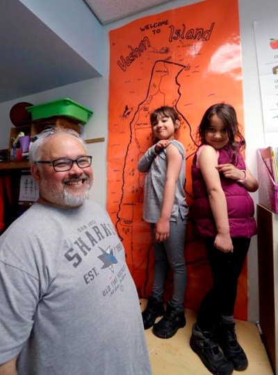 In this photo taken Wednesday, May 15, 2019, Gator Lanphear smiles as his twin six-year-old daughters Scarlett, center, and Leilani show off where on their arms they received their measles immunizations while posing for a photo in Vashon Island, Wash. Lanphear said he not only immunizes his twin daughters but instills in them the importance of getting their shots as a heroic act. The number of philosophical exemptions in Vashon has decreased dramatically, but overall, there are still 11.6 percent of students whose families elect not to vaccinate. (AP Photo/Elaine Thompson)