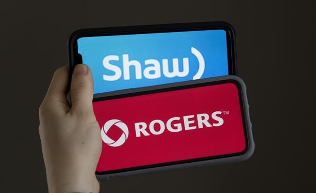 A woman holds two cellphones with the corporate logos of Rogers Communications and Shaw Communications in a photo illustration in Chelsea, Que., Monday March 29, 2021. A review of the proposed $26-billion purchase of Shaw Communications by Rogers Communications continues into its third day today in Ottawa before a committee of federal MPs. THE CANADIAN PRESS/Adrian Wyld