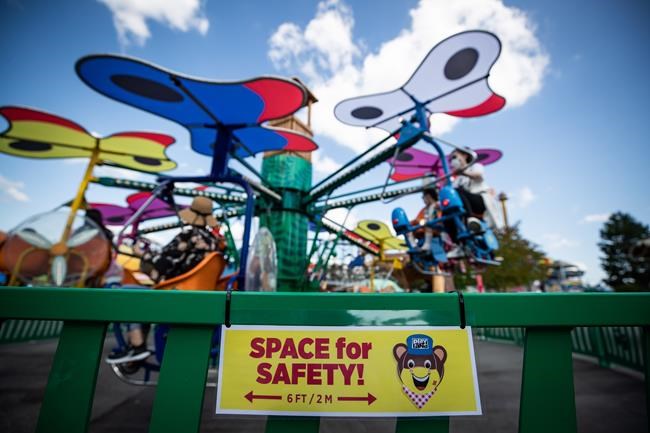 A sign reminding guests to stay 2 metres apart is seen on a fence as people ride an attraction at Playland amusement park at the Pacific National Exhibition, in Vancouver, on Friday, July 10, 2020. A Vancouver amusement park has delayed it opening until after the May long weekend amid concerns it will attract COVID-19 rule breakers. THE CANADIAN PRESS/Darryl Dyck