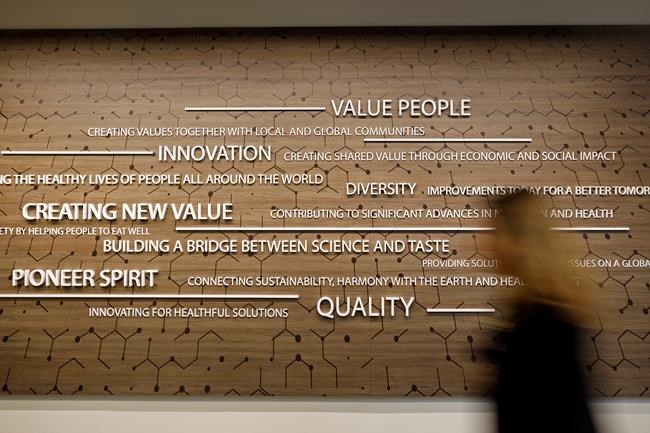 An employee of Ajinomoto, a global food and pharmaceutical company, walks past the company motto in Itasca, Ill., Monday, June 7, 2021. In May, Ajinomoto's employees returned to in-person work in a space designed for a post-COVID world. Hallways are wider. Glass panels separate cubicles. A work space has been transformed into a spa-like relaxation area. The culinary center is wired for virtual presentations. And a cleaning crew comes through twice a day, leaving Post-it notes to show what's been disinfected. (AP Photo/Shafkat Anowar)