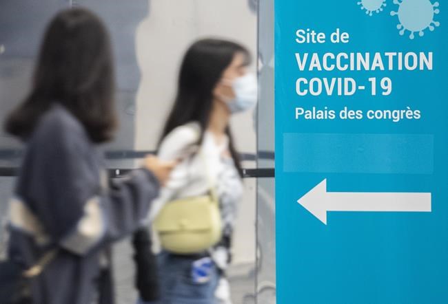 People wear face masks as they walk by a COVID-19 vaccination sign in Montreal, Sunday, Oct. 10, 2021, as the COVID-19 pandemic continues in Canada and around the world. THE CANADIAN PRESS/Graham Hughes
