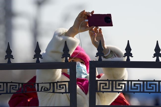 A woman outside the fenced off closed-loop area for the Olympics takes a picture of the Olympic Green ahead of the 2022 Winter Olympics, Feb. 1, 2022, in Beijing. (AP Photo/Natacha Pisarenko)