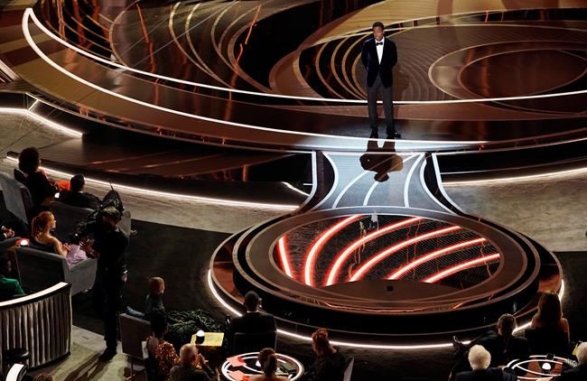 Presenter Chris Rock speaks onstage as Will Smith and Jada Pinkett, bottom left, listen from their seats at the Oscars, Sunday, March 27, 2022, at the Dolby Theatre in Los Angeles. (AP Photo/Chris Pizzello)
