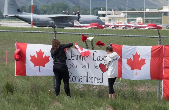 The Canadian Forces Snowbirds jets are seen in the background as women attach a sign to a fence in Kamloops, B.C., Monday, May 18, 2020. Capt. Jenn Casey died Sunday after the Snowbirds jet she was in crashed shortly after takeoff. The pilot of the aircraft is in hospital with serious injuries. THE CANADIAN PRESS/Jonathan Hayward