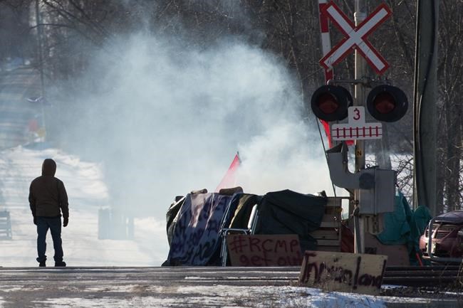 A protester stands beside smoke at the closed train tracks in Tyendinaga Mohawk Territory near Belleville, Ont., on Thursday Feb. 20, 2020, as they protest in solidarity with the Wet'suwet'en hereditary chiefs opposed to the LNG pipeline in northern British Columbia. A group of hereditary leaders from the Wet'suwet'en First Nation in B.C. is to spend the day with Mohawk supporters in Ontario. THE CANADIAN PRESS/Lars Hagberg