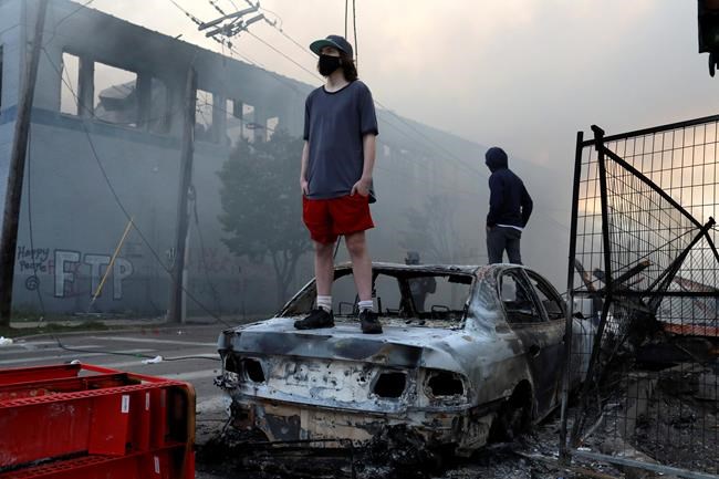 People stand on a burned up car as fires burn near a Target Store after a night of unrest and protests in the death of George Floyd early Thursday, May 28, 2020 in downtown Minneapolis. Floyd died after being restrained by Minneapolis police officers on Memorial Day. A video taken by a bystander shows a Minneapolis police officer with his knee on the neck of a man in custody who later died. The four officers involved have been fired. (David Joles/Star Tribune via AP)