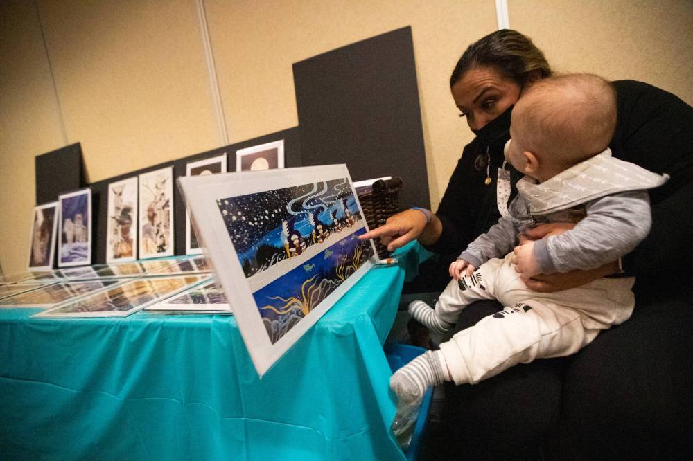 Renata Meconse shows her nephew, Thomas Thompson, prints of little people at the Ziigwan (Spring) Indigenous Arts Market & Tradeshow at Canad Inns Polo Park Saturday. (Daniel Crump / Winnipeg Free Press)