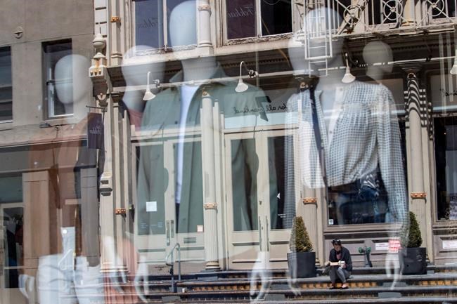 FILE - In this May 7, 2020 file photo a woman sitting on a stoop reading a book in the sun is seen reflected by a closed clothing store's window on West Broadway in the SoHo neighborhood of the Manhattan borough in New York. U.S. retail sales likely recovered somewhat in May as the economy increasingly reopened from closures caused by the coronavirus and more shoppers felt confident enough to spend. The Commerce Department is expected to report Tuesday, June 16, 2020 that retail purchases climbed 9% from April to May, according to economists surveyed by the data provider FactSet. (AP Photo/Mary Altaffer, file)