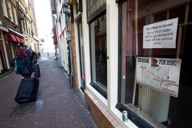 Two tourists pull their suitcases passing closed windows of sex workers in the Red Light District of Amsterdam, Netherlands, Monday, March 16, 2020. Health minister Bruno Bruins ordered all Dutch schools, cafes, restaurants coffeeshops, saunas, sex clubs and sport and fitness clubs to be closed from Sunday March 15th onwards as the government sought to prevent the further spread of coronavirus in the Netherlands. For most people, the new coronavirus causes only mild or moderate symptoms, such as fever and cough. For some, especially older adults and people with existing health problems, it can cause more severe illness, including pneumonia. (AP Photo/Peter Dejong)