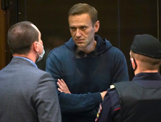 In this handout photo provided by Moscow City Court Russian opposition leader Alexei Navalny talks to one of his lawyers, left, while standing in the cage during a hearing to a motion from the Russian prison service to convert the suspended sentence of Navalny from the 2014 criminal conviction into a real prison term in the Moscow City Court in Moscow, Russia, Tuesday, Feb. 2, 2021. (Moscow City Court via AP)