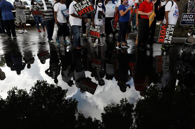 People carrying signs supporting voting rights are reflected in a puddle as they arrive at an early voting center at Model City Branch Library, as part of a