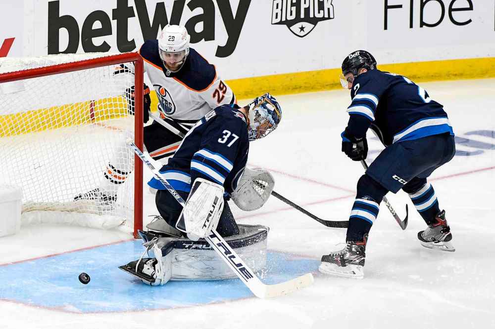 Leon Draisaitl shoots the puck through the crease by goaltender Connor Hellebuyck during the first overtime period. (Fred Greenslade / The Canadian Press)