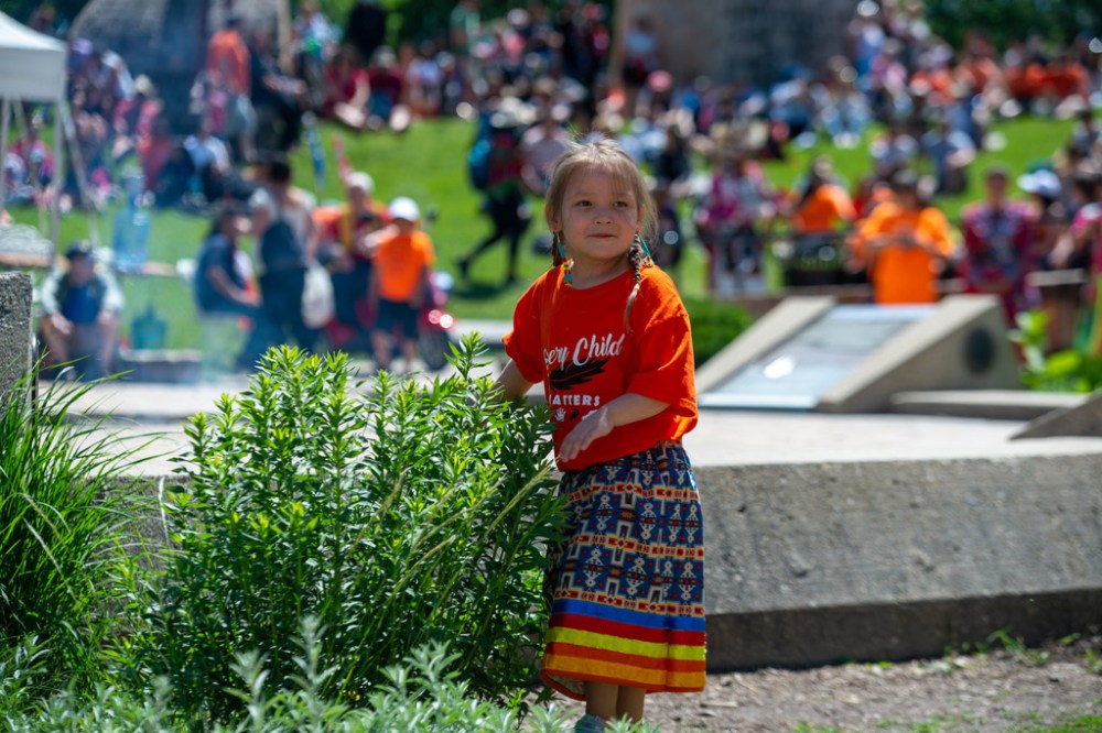 ETHAN CAIRNS / WINNIPEG FREE PRESS
A child dressed a traditional outfit with an orange shirt play with a plant before the pow-wow dance demonstration at A New Day Ceremony at The Forks in Winnipeg, Manitoba on Friday, July 1, 2022