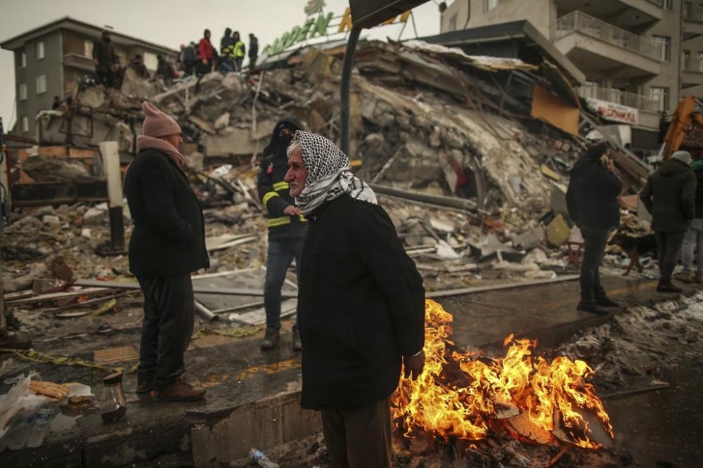 People warm themselves next to a collapsed building in Malatya, Turkey, Tuesday, Feb. 7, 2023. Search teams and aid are pouring into Turkey and Syria as rescuers working in freezing temperatures dig through the remains of buildings flattened by a magnitude 7.8 earthquake. (AP Photo/Emrah Gurel)