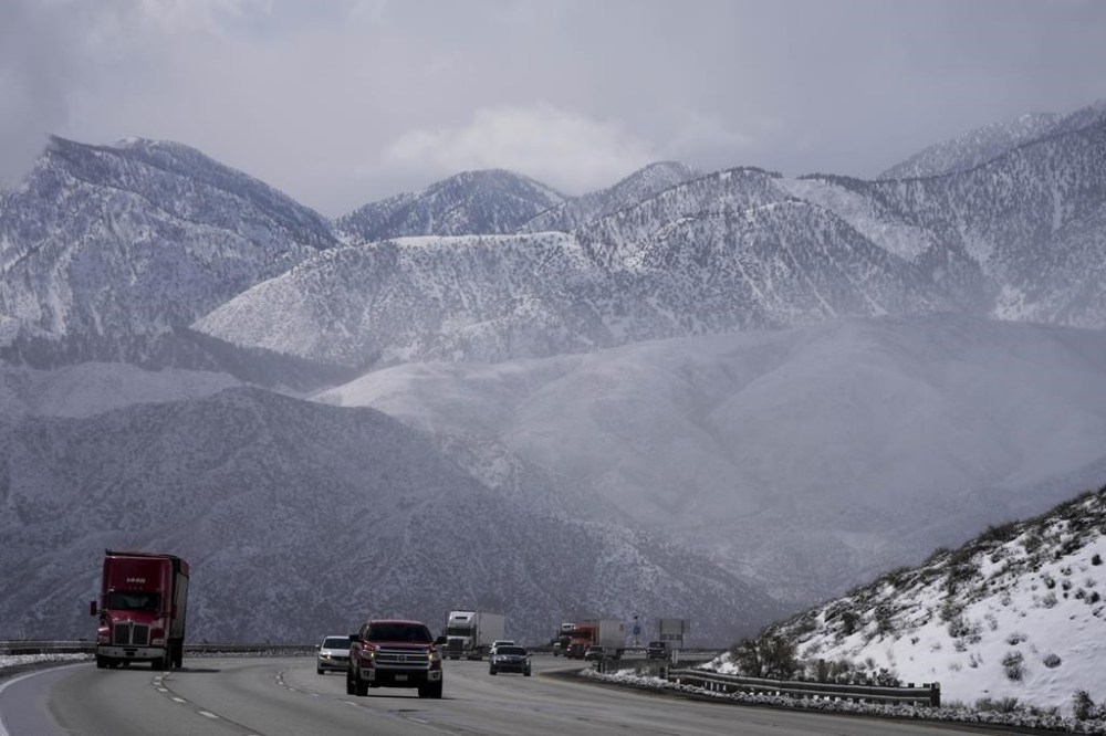 Vehicles make their way along the I-15 as clouds pass through the snow-covered mountains near Hesperia, Calif., Wednesday, March 1, 2023. Emergency crews are scrambling to shuttle food and medicine to residents of California mountain communities stranded by back-to-back winter storms. (AP Photo/Jae C. Hong)