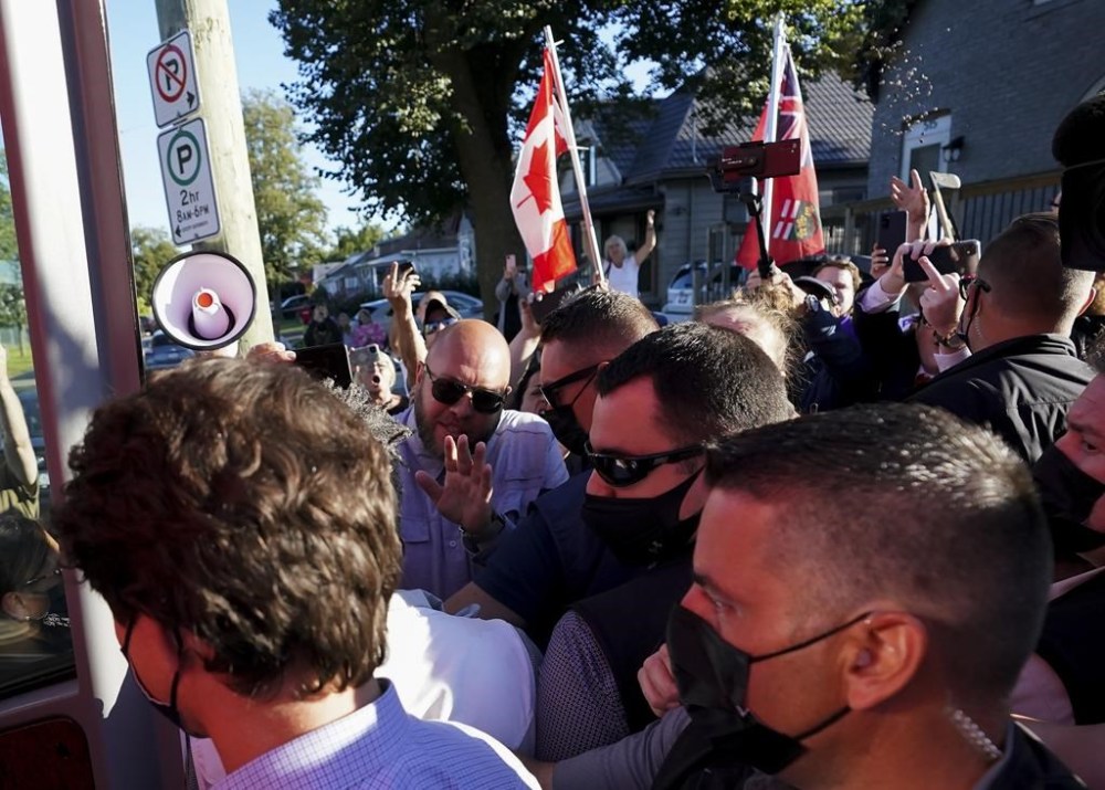 A man, top right, throws gravel at Liberal leader Justin Trudeau, left, as the RCMP security detail provide protection at a local microbrewery during the Canadian federal election campaign in London Ont., on Monday, September 6, 2021. An Ontario man accused of throwing gravel at Justin Trudeau during a September 2021 campaign stop has pleaded guilty to a charge of common assault. THE CANADIAN PRESS/Nathan Denette