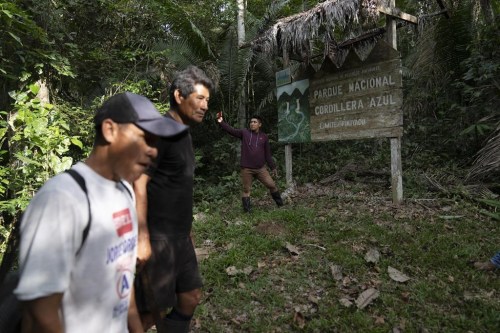 A group of men pass in front of a sign announcing the limit of Cordillera Azul National Park in Peru's Amazon, Monday, Oct. 3, 2022. Analysis by independent experts and reporting by The Associated Press raise doubts about whether a program to sell carbon credits is delivering on its promise to stop deforestation in the park and balance out carbon emissions by the companies buying the credits. (AP Photo/Martin Mejia)