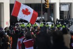 A person waves a Canadian flag as police on horseback and an armoured police vehicle are positioned in front of protesters, on the 22nd day of a protest against COVID-19 measures that has grown into a broader anti-government protest, in Ottawa, on Friday, Feb. 18, 2022. Lawyers for the federal attorney general are set to expand today on the legal rationale for the historic use of the Emergencies Act to dispel 