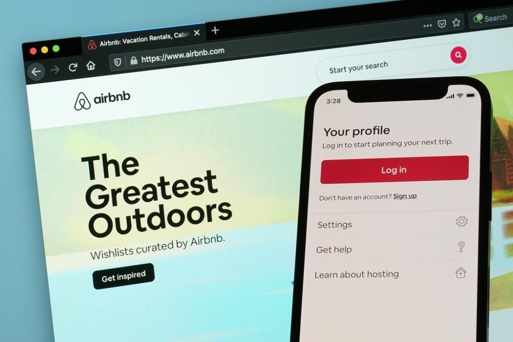 FILE - The login page for Airbnb's iPhone app is seen in front of a computer displaying Airbnb's website on May 8, 2021, in Washington. Airbnb is going back to its roots to cater to travelers who are growing more concerned with saving money. The company said Wednesday, May 3, 2023, that it's rolling out a new Airbnb Rooms offering in which guests share space with the owner. Airbnb says room prices will average $67 a night. (AP Photo/Patrick Semansky, File)