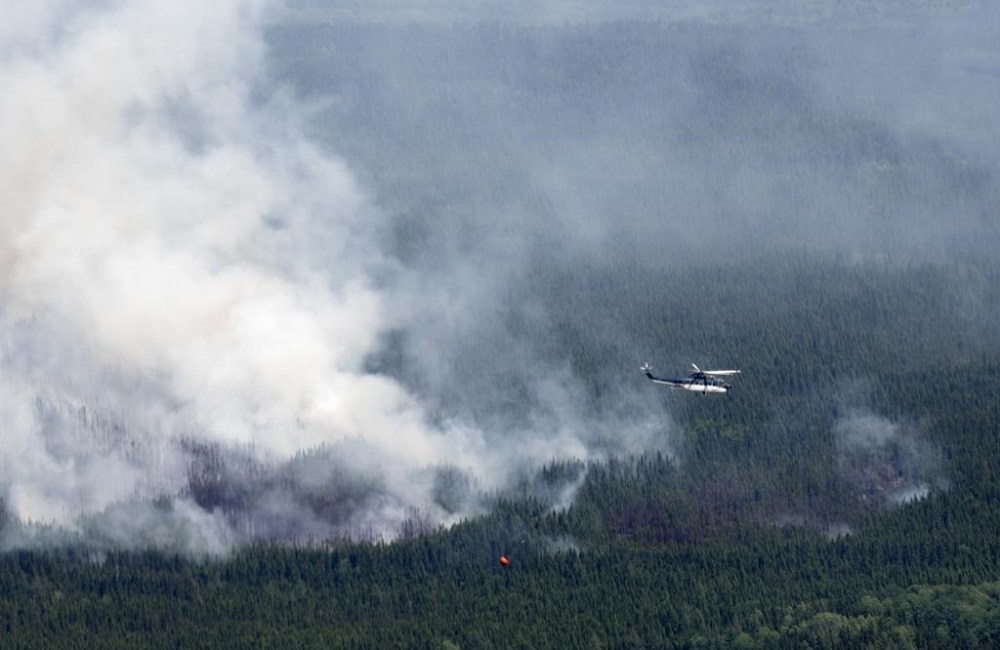 A helicopter carrying a water basket flies past a smoke plume near Lebel-sur-Quevillon, Que., Wednesday, July 5, 2023. Natural Resources Canada says the country's record-breaking wildfire season will continue to be greater than normal throughout July and into August. THE CANADIAN PRESS/Adrian Wyld