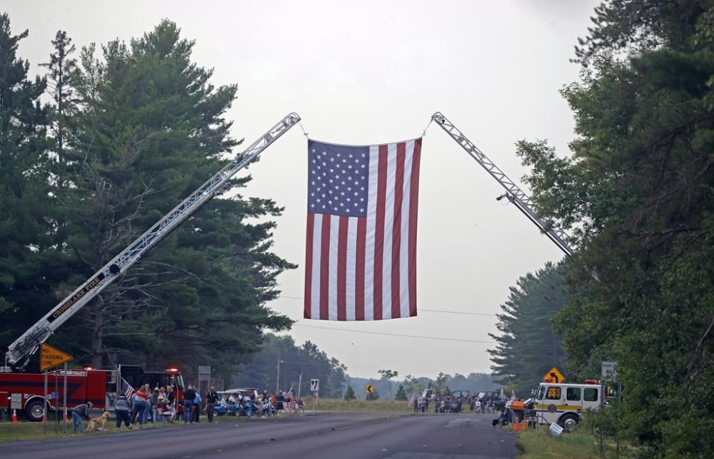 A large flag hangs over Patriot Avenue for the funeral services for Fargo Police Officer Jake Wallin in Pequot Lakes, Minn., on Saturday, July 22, 2023. Wallin, 23, was killed July 14 when a man armed with 1,800 rounds of ammunition, multiple guns and explosives ambushed officers responding to a routine traffic crash. (David Samson/Forum Communications Co. via AP, POOL)