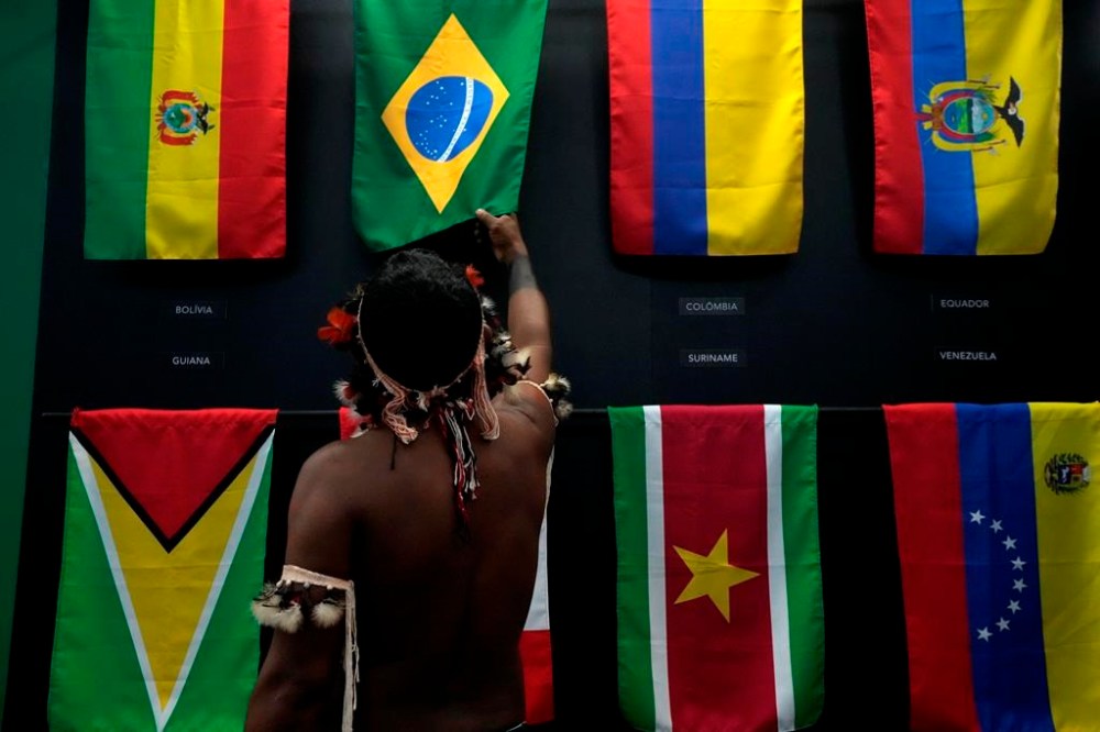 FILE - An indigenous man passes his hand over the Brazilian flag in a flag pavilion of the States parties to the Amazon Cooperation Treaty, during the Amazon Dialogue meetings at the Hangar convention center in Belem, Brazil, Aug. 6, 2023. The two-day Amazon Summit opens Aug. 8, 2023, in Belem, where Brazil hosts policymakers and others to discuss how to tackle the immense challenges of protecting the Amazon and stemming the worst of climate change. (AP Photo/Eraldo Peres, File)