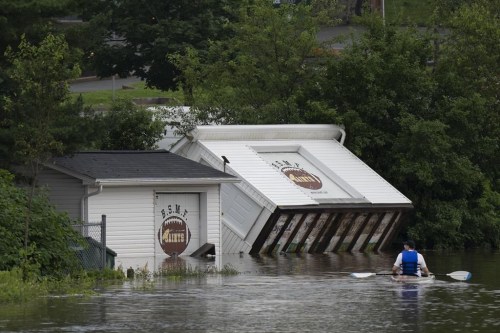 Buildings are seen upended on a riverbank as a man paddles a kayak through floodwater following a major rain event in Halifax on Saturday, July 22, 2023. Nova Scotia's premier is mulling letting small towns issue emergency alerts during flooding, but one expert says there are wider problems behind the province's faulty response. THE CANADIAN PRESS/Darren Calabrese