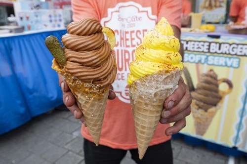 An employee of the So Cute Ice Cream stall holds up a cheese-coated waffle cone, topped with a pretzel and fresh pickle, left, and a Street Corn Ice Cream with lime, cotija cheese, and chilli seasoning during a Canadian National Exhibition (CNE) media preview in Toronto on Wednesday, Aug. 16, 2023. THE CANADIAN PRESS/Spencer Colby