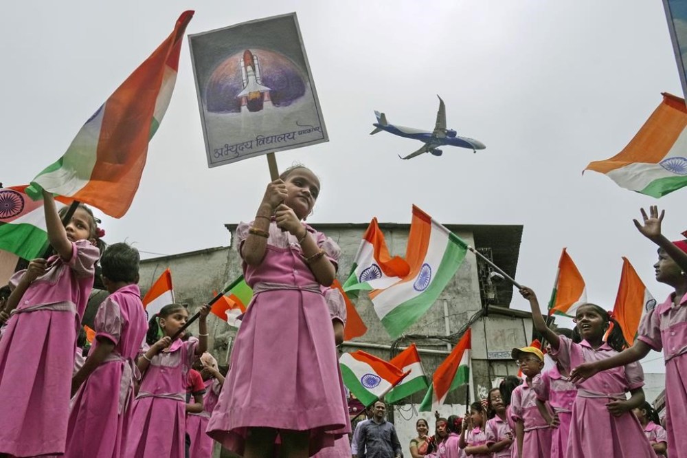 An aircraft prepares to land as children cheering for the successful landing of India’s moon craft Chandrayaan-3, on the moon surface, wave Indian flags at a school near the airport in Mumbai, India, Tuesday, Aug.22, 2023. India’s previous attempt to land a robotic spacecraft near the moon’s little-explored south pole ended in failure in 2019. (AP Photo/ Rajanish Kakade)