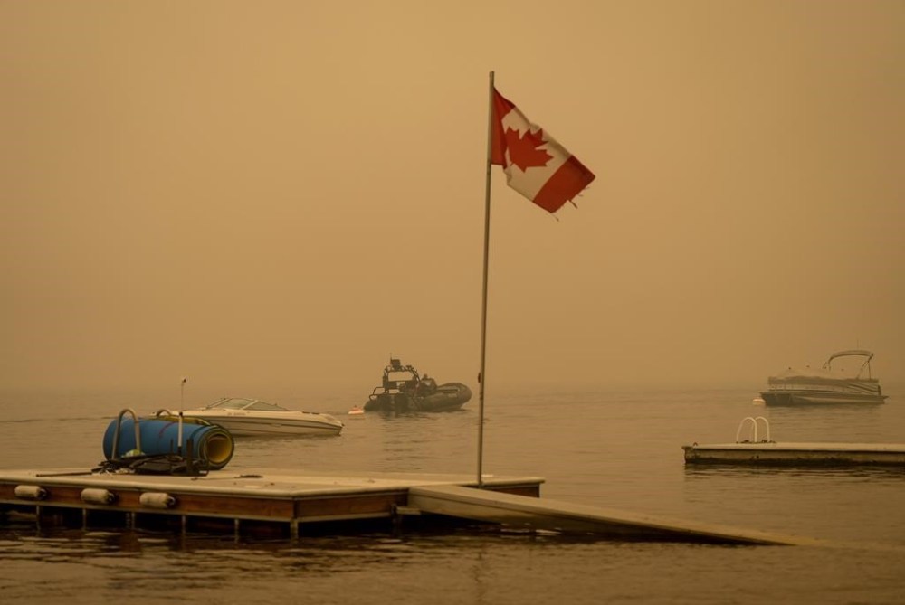 Thick smoke from the Lower East Adams Lake wildfire fills the air and a Canadian flag flies in the wind as RCMP officers on a boat patrol Shuswap Lake, in Scotch Creek, B.C., on Sunday, August 20, 2023. THE CANADIAN PRESS/Darryl Dyck