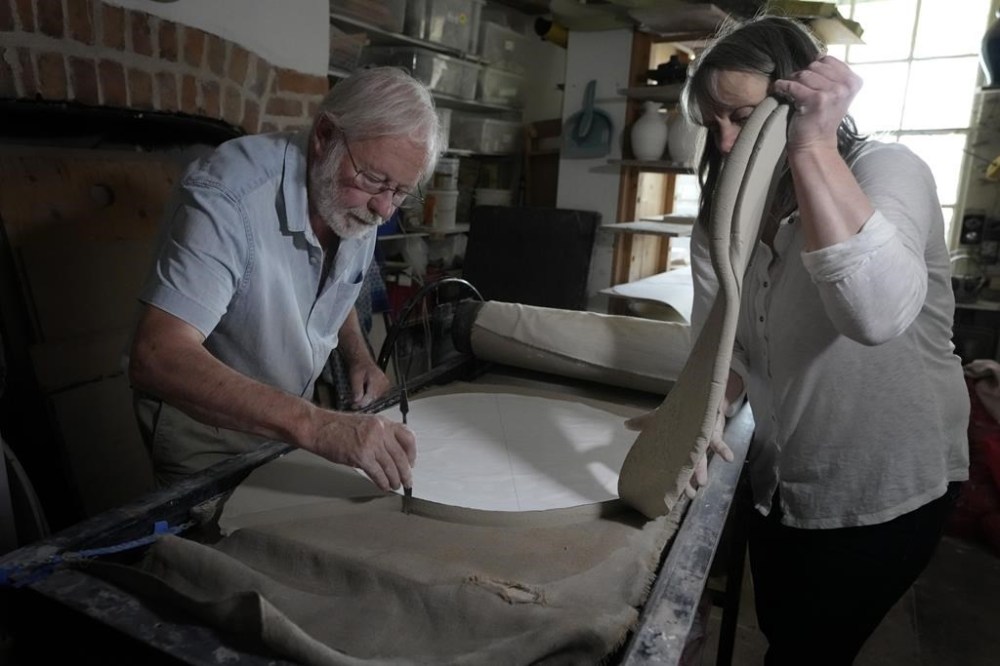 Ned Heywood cuts out the initial shape from a rolled slab of clay as Julia Land takes away the excess during the making of an English Heritage Blue Plaque, at Heritage Ceramics, The Workshop Gallery in Chepstow, Wales Wednesday, Sept. 6, 2023. English Heritage is preparing to unveil its 1,000th blue plaque, the famous discs that dot the walls of buildings throughout London, marking the places where scientists, artists, politicians and activists have made history. The charity is working to broaden the program to include more women, people from minority ethnic backgrounds and community groups so that it better reflects the diversity of the capital. (AP Photo/Kirsty Wigglesworth)
