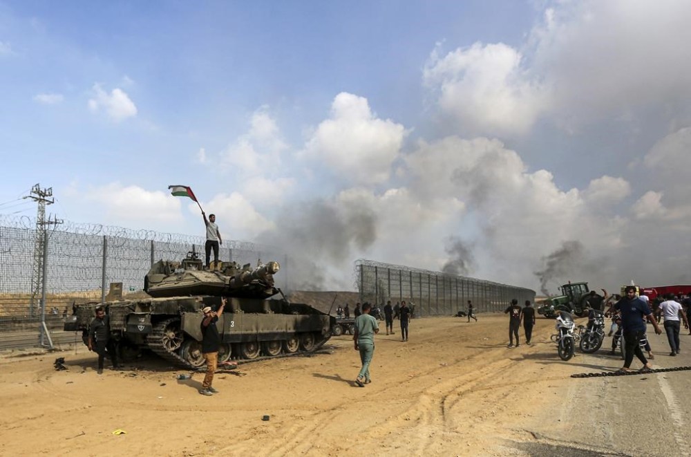 Palestinians wave their national flag and celebrate by a destroyed Israeli tank at the Gaza Strip fence east of Khan Younis southern Saturday, Oct. 7, 2023. The militant Hamas rulers of the Gaza Strip carried out an unprecedented, multi-front attack on Israel at daybreak Saturday, firing thousands of rockets as dozens of Hamas fighters infiltrated the heavily fortified border in several locations by air, land, and sea and catching the country off-guard on a major holiday. (AP Photo/Yousef Masoud)
