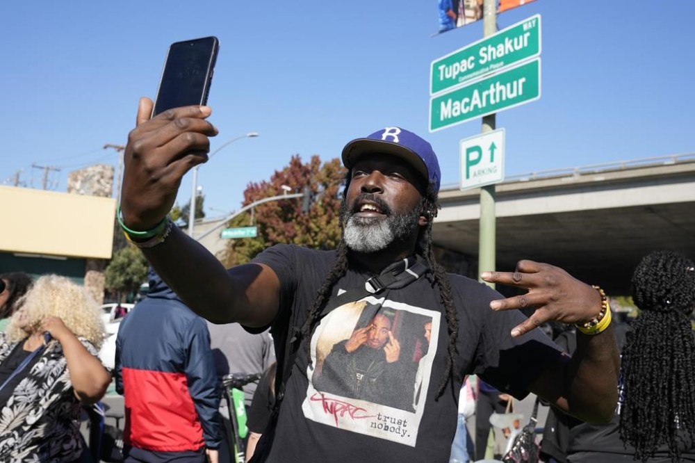 Randy Moore, of Los Angeles, poses beneath a new sign during a street renaming ceremony for Tupac Shakur in Oakland, Calif., Friday, Nov. 3, 2023. A stretch of street in Oakland was renamed for Shakur, 27 years after the killing of the hip-hop luminary. A section of Macarthur Boulevard near where he lived in the 1990s is now Tupac Shakur Way, after a ceremony that included his family members and Oakland native MC Hammer. (AP Photo/Eric Risberg)