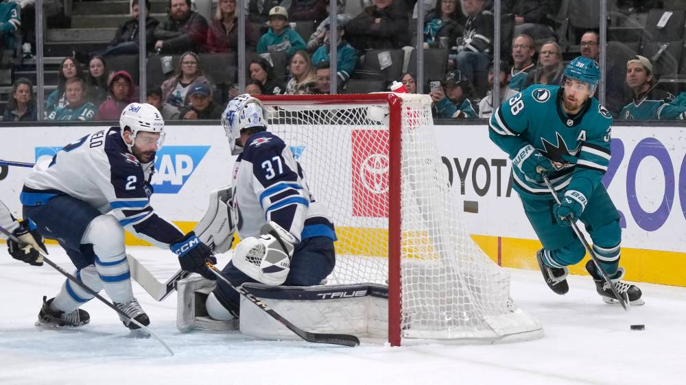 San Jose Sharks defenceman Mario Ferraro carries the puck behind the net, as Winnipeg Jets defenceman Dylan DeMelo and goaltender Connor Hellebuyck keep an eye out for a pass during the first period. (AP Photo/Jeff Chiu)