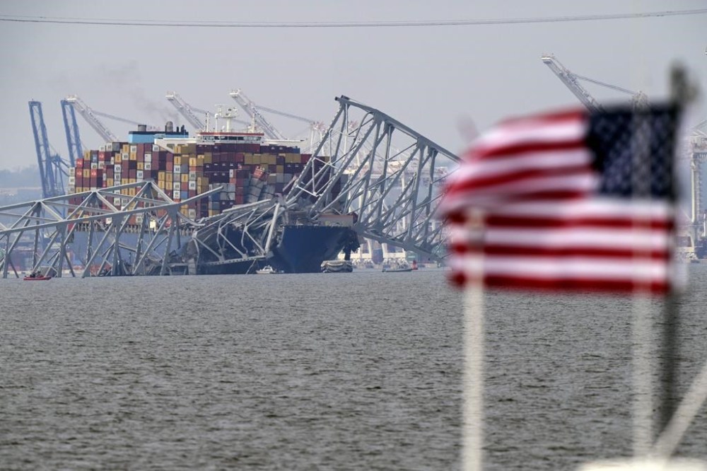 An American flag flies on a moored boat as a container ship rests against wreckage of the Francis Scott Key Bridge on Tuesday, March 26, 2024, as seen from Pasadena, Md. (AP Photo/Mark Schiefelbein)
