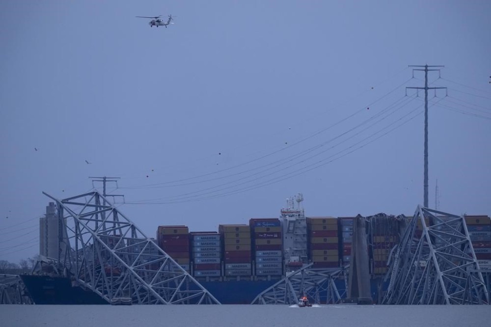 A helicopter flies overhead as a container ship rests against wreckage of the Francis Scott Key Bridge as night falls on Tuesday, March 26, 2024, as seen from Sparrows Point, Md. The ship rammed into the major bridge in Baltimore early Tuesday, causing it to collapse in a matter of seconds and creating a terrifying scene as several vehicles plunged into the chilly river below. (AP Photo/Matt Rourke)