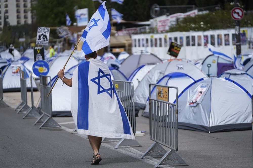 A man walks with the Israeli flag past a camp set up by people who are protesting against Israeli Prime Minister Benjamin Netanyahu's government and call for the release of hostages held in the Gaza Strip by the Hamas militant group near the Knesset, Israel's parliament, in Jerusalem, Tuesday, April 2, 2024. (AP Photo/Ohad Zwigenberg)