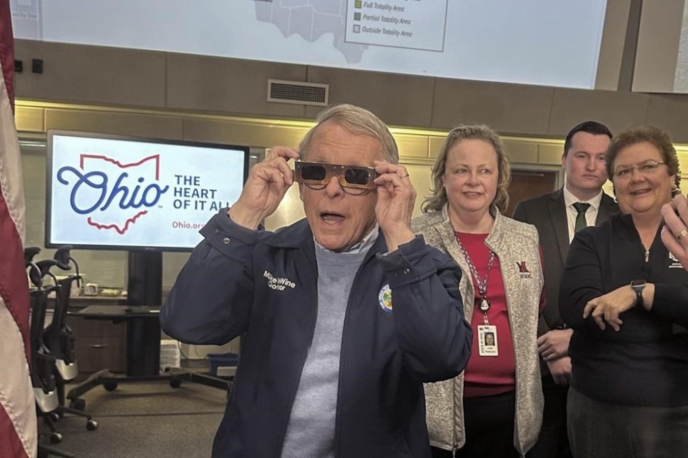 Republican Ohio Gov. Mike DeWine sports a pair of eclipse glasses while talking to reporters at the Ohio Emergency Operations Center in Columbus, Ohio, on Friday, April 5, 2024. DeWine activated the center on Friday to assist communities before, during and after Monday's total solar eclipse, which could draw hundreds of thousands of tourists to the state. (AP Photo/Julie Carr Smyth)