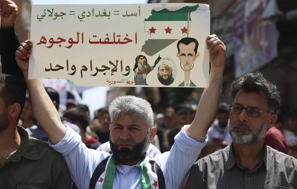 FILE - A Syrian protester holds up a placard during a protest against al-Qaida-linked Hayat Tahrir al-Sham group in Binnish town, in Idlib province, Syria, on May 17, 2024. Syria's devastating civil war, now in its 14th year, remains largely frozen and so are efforts to find a viable political solution to end it, while millions of Syrians have been pulled into poverty, and struggle with accessing food and health care as the economy deteriorates across the country's front lines. Aid organizations are making their annual pitches to donors ahead of a fundraising conference in Brussels for Syria on Monday, May 27. The placard shows drawings of Syrian President Bashar Assad, the late Islamic State group leader Abu Bakr al-Baghdadi and Abu Mohammed al-Golani, leader of the Hayat Tahrir al-Sham, once linked to al-Qaida. The Arabic writing reads: