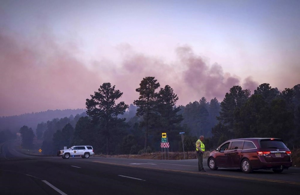Smoke from the South Fork Fire reflects a pink hue during sunrise Tuesday, June 18, 2024 as law enforcement officers turn drivers away on State Road 48 at State 220 because of wildfires burning near Ruidoso, N.M. The South Fork Fire has consumed nearly 14,000 acres. (Chancey Bush/The Albuquerque Journal via AP)
