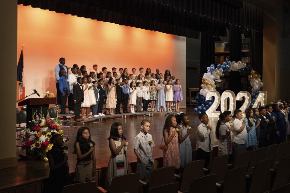 Students sing the national anthem during a graduation ceremony at Trevor Day School, Friday, June 21, 2024 in New York. Thousands of migrant families in New York City are facing a summer of uncertainty for their school-aged children with a citywide limit of 60 days in a shelter before needing to reapply or find their own. (AP Photo/Jeenah Moon)