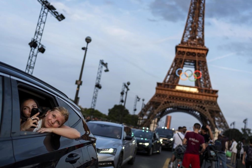 FILE - Passengers in the back of a taxi film themselves as they leave the Eiffel Tower decorated with the Olympic rings ahead of the 2024 Summer Olympics, Wednesday, July 17, 2024, in Paris. The city itself will be one of the protagonists of the 2024 Olympics. That's why the Eiffel Tower and Seine River are featured just as prominently as Simone Biles or Katie Ledecky in NBC’s ads. (AP Photo/David Goldman, File)