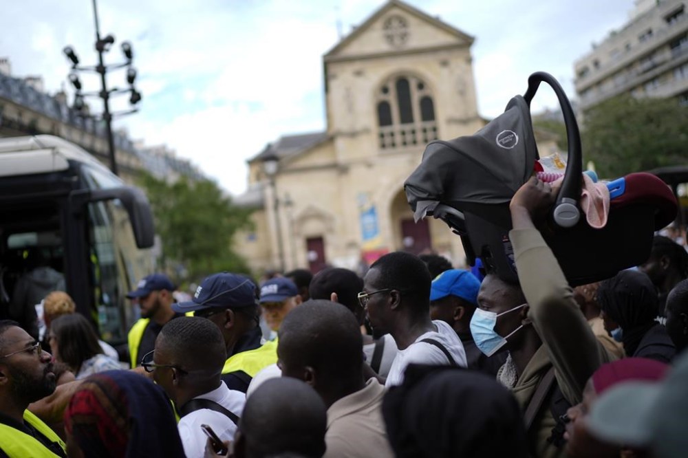 A man carries a baby carrier before boarding a police after a migrants' protest at the 2024 Summer Olympics, Thursday, July 25, 2024, in Paris, France. On the eve of the grandiose opening ceremony for the Paris Olympics, police cleared out migrants sleeping in a tent camp in the capital as social and environmental advocacy groups raise attention to criticisms of the Paris Games such as the displacement of migrants and housing issues. (AP Photo/David Goldman)