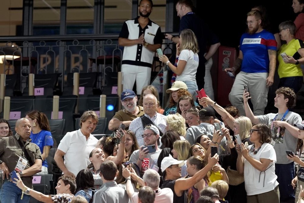 Actor Tom Cruise, lower left in white shirt, and entertainer John Legend, top center, in white and brown shirt, take photos with fans as they attend the women's artistic gymnastics qualification round at the 2024 Summer Olympics, Sunday, July 28, 2024, in Paris, France. (AP Photo/Abbie Parr)